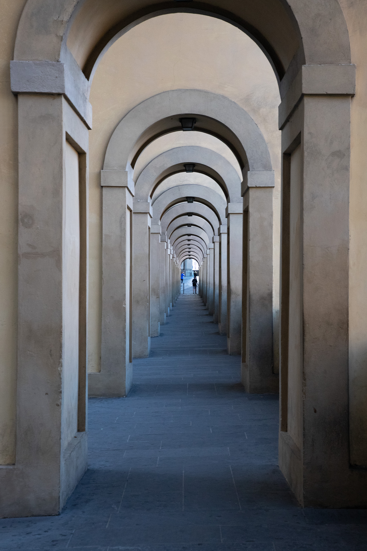 View through a series of limestone arches leading to a distant street with people walking.