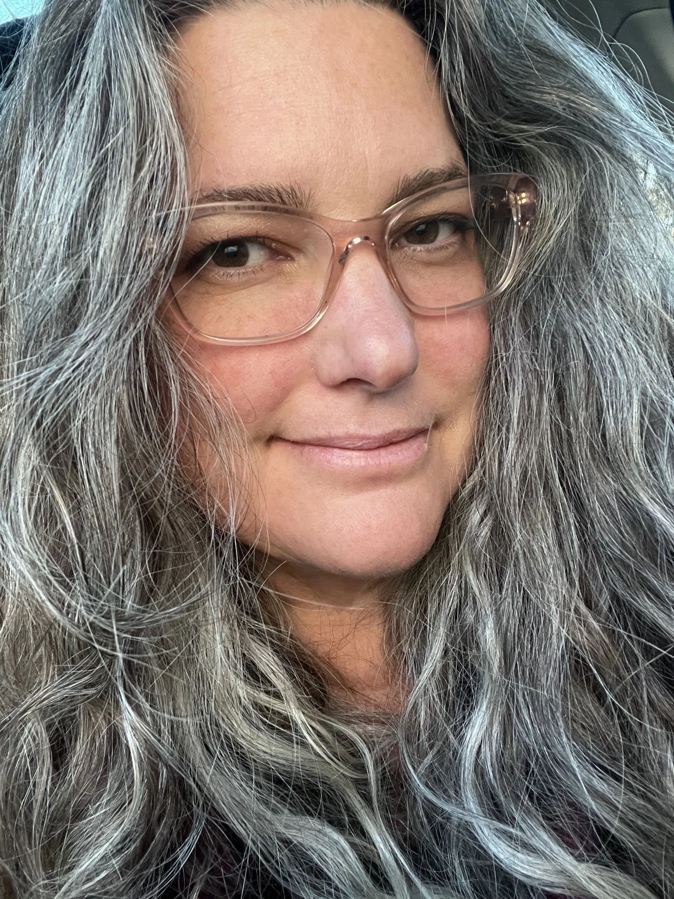 Close-up of a woman with long, wavy gray and black hair, wearing clear glasses, with a subtle smile on her face.