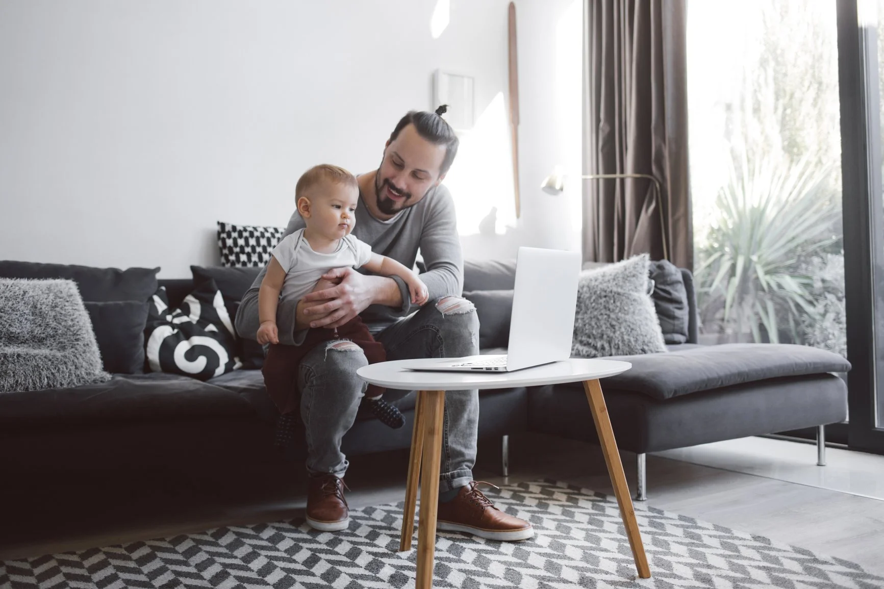 a new father smiling at his baby while holding him and sitting in front of an open laptop