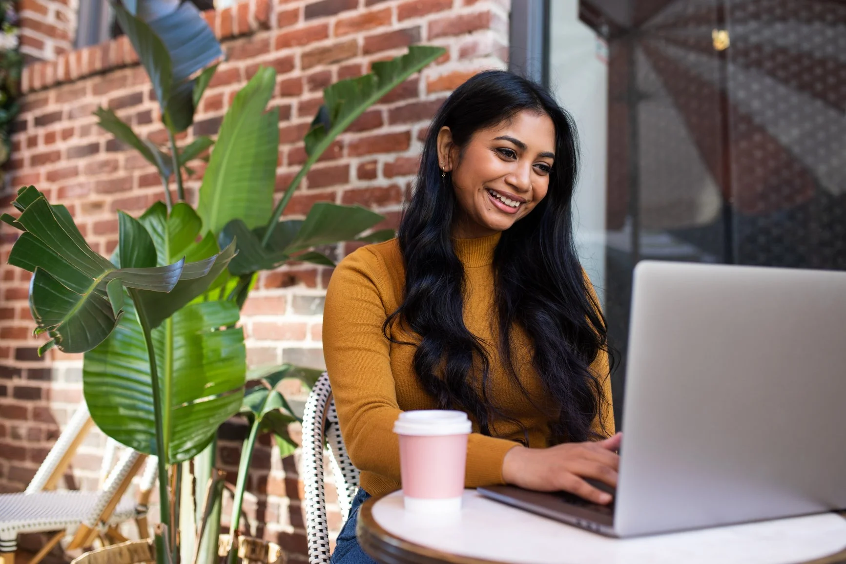 a woman of color smiling while working on her laptop outside of a brick building with to-go cup of coffee close by