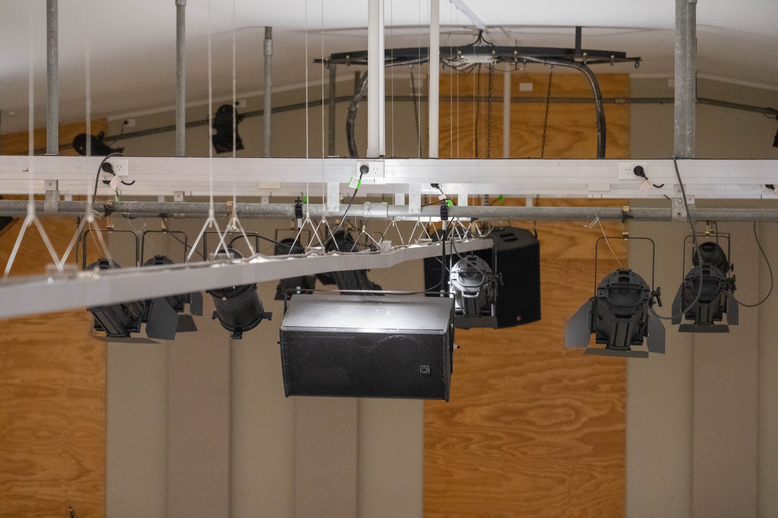 Ceiling-mounted stage lighting system with multiple black spotlights and a black speaker, connected by electrical cables, in a room with wooden walls.