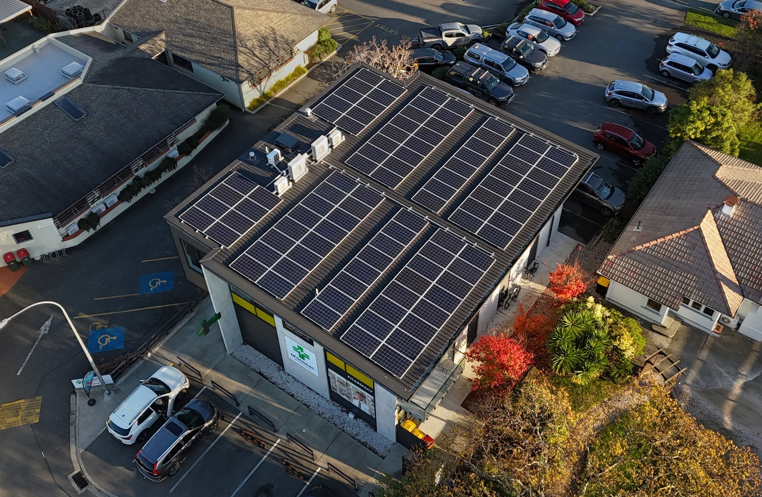 An aerial view of a city block showing a pharmacy with solar panels on its roof, surrounded by parked cars, trees, and adjacent buildings.