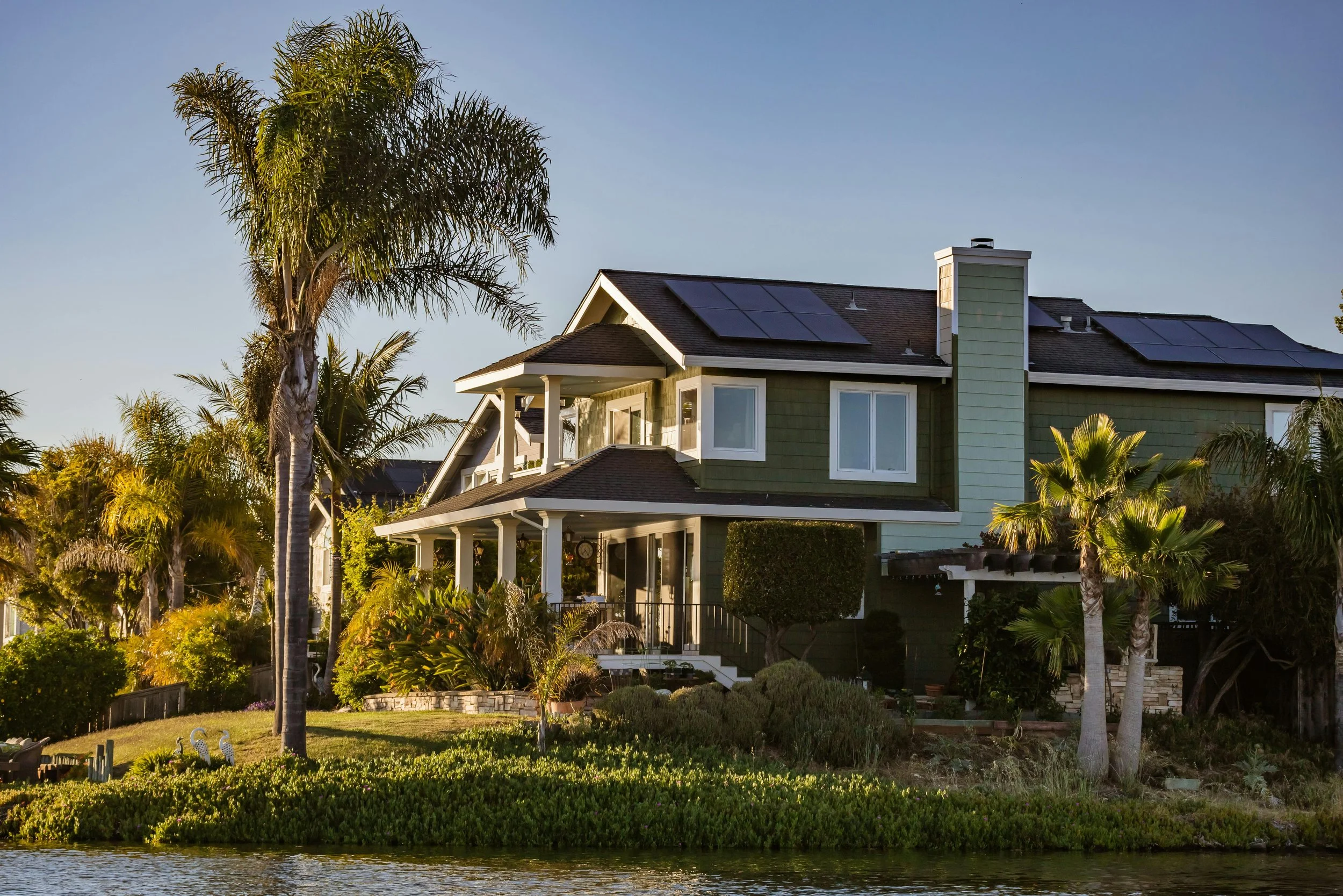 A house with solar panels on the roof surrounded by palm trees and lush greenery, with a body of water in the foreground.