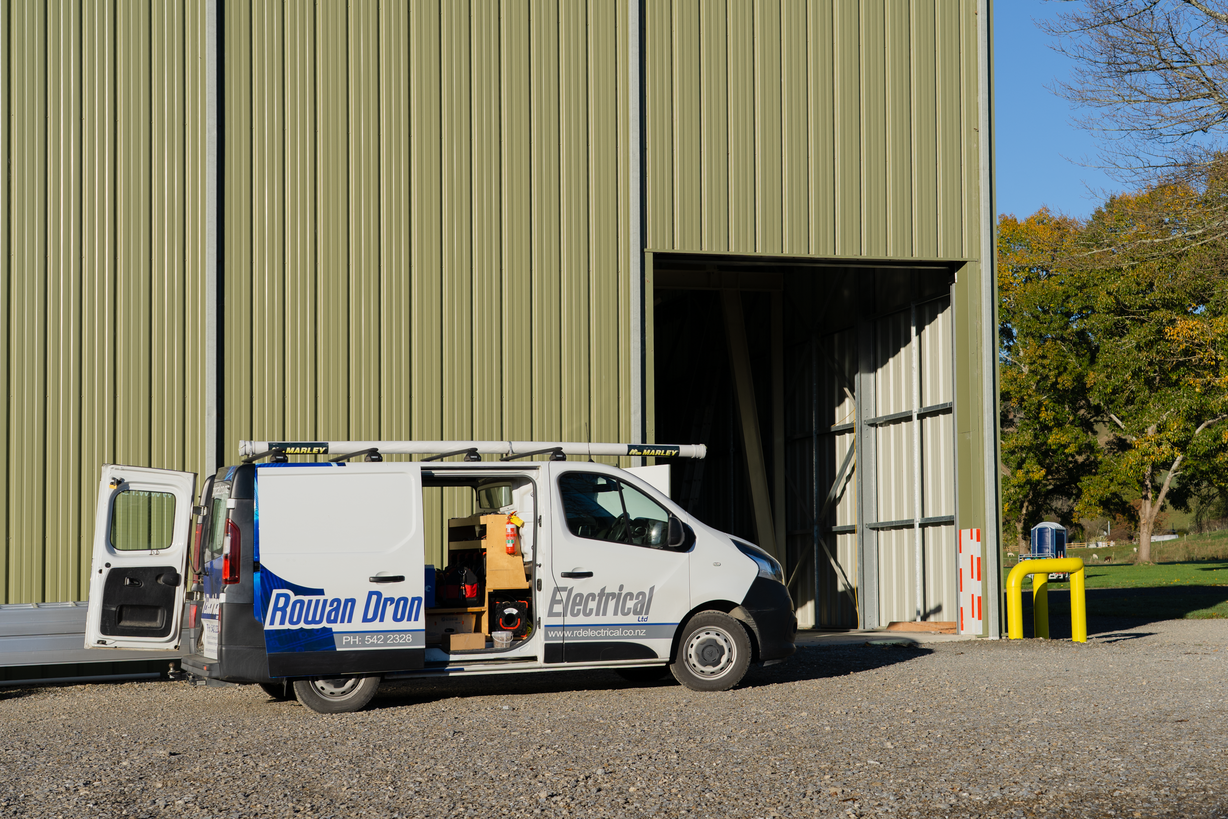 A white service van with open side door parked in front of a green metal building under a clear blue sky. The van displays the logo ‘Rowan Dron Electrical’ and includes contact details. Construction materials and tools are inside the van.
