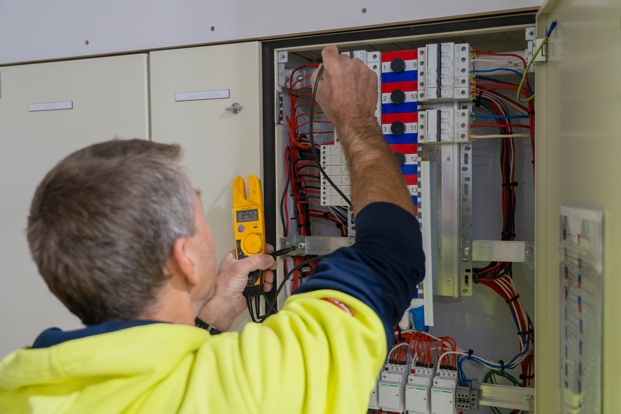 An electrician in a yellow safety vest using a multimeter to test electrical wiring inside an electrical panel or control box.