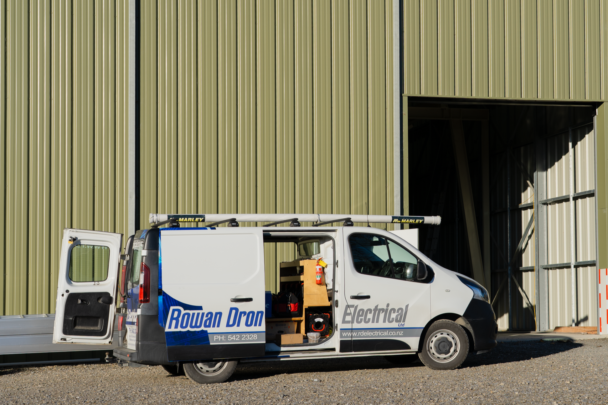 A white service van with Rowan Dron Electrical branding parked in front of a large industrial building with green metal siding. The van's side door is open, revealing tools and equipment inside.