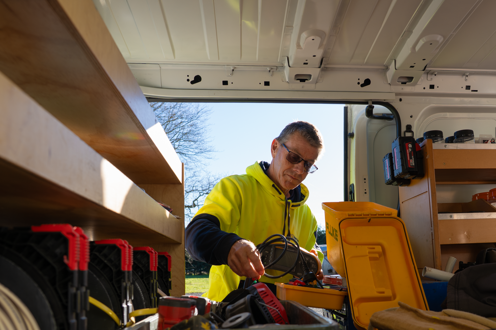 An electrician in safety glasses and a yellow jacket working with tools in the back of a work van, with shelves filled with containers and tools inside the van and a clear blue sky outside.