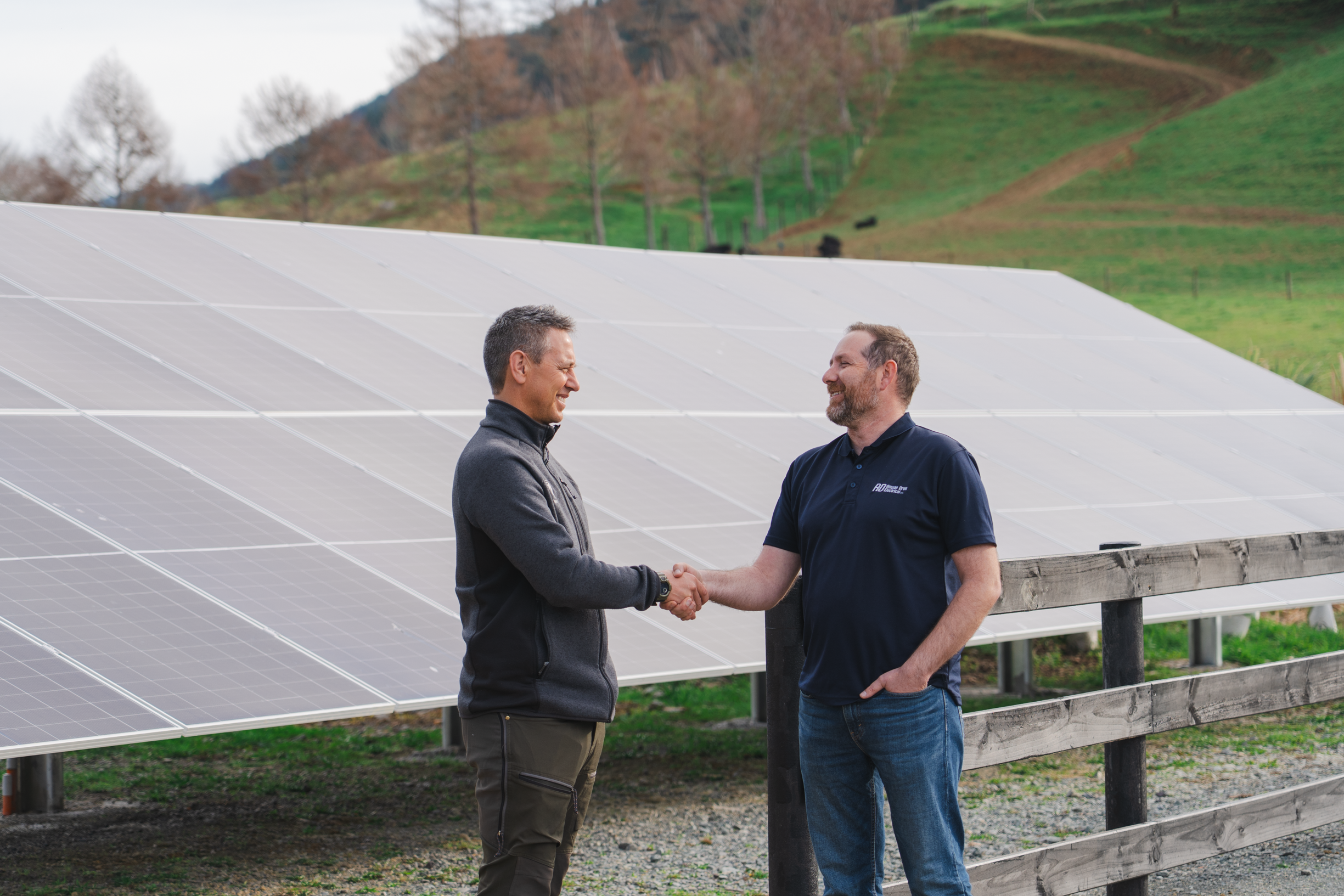 Rowan and a happy client shaking hands in front of solar panels on a farm with hills and trees in the background.