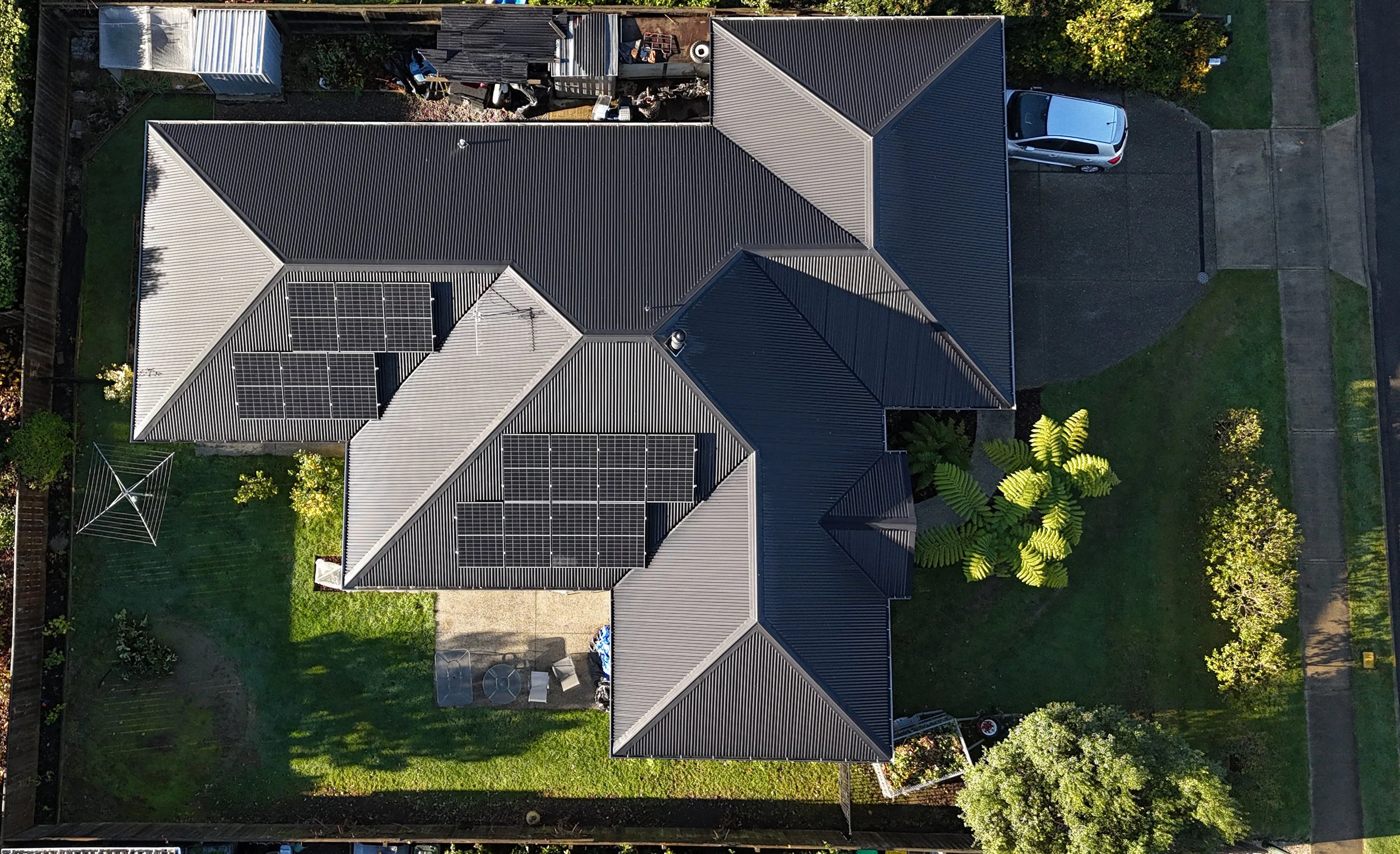 Aerial view of a house with a metal roof, solar panels, a driveway with a car, surrounded by a backyard with a clothesline, trees, and garden decorations.