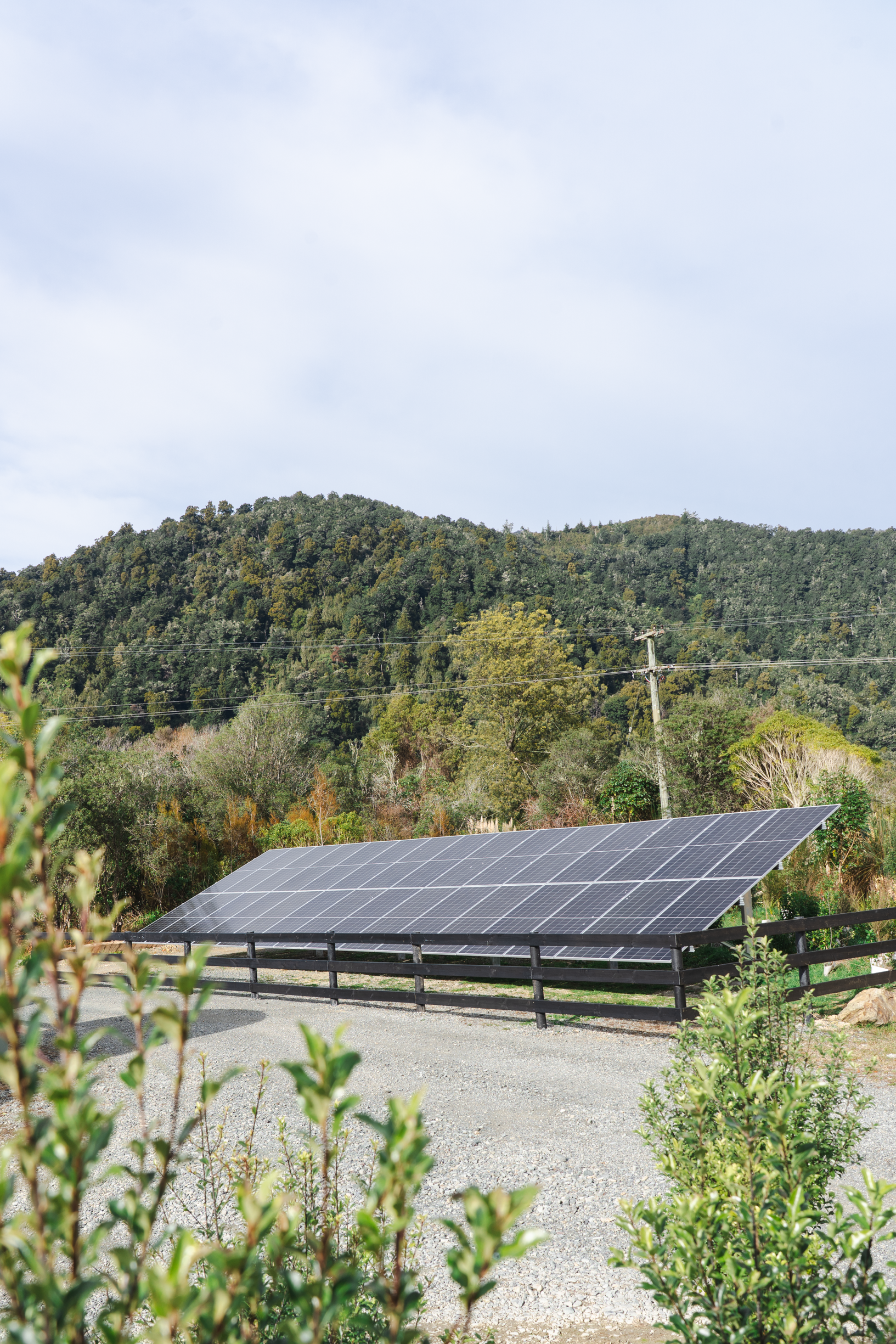 Solar panels installed on a fenced area in a rural landscape with a wooded hill in the background, under a partly cloudy sky.