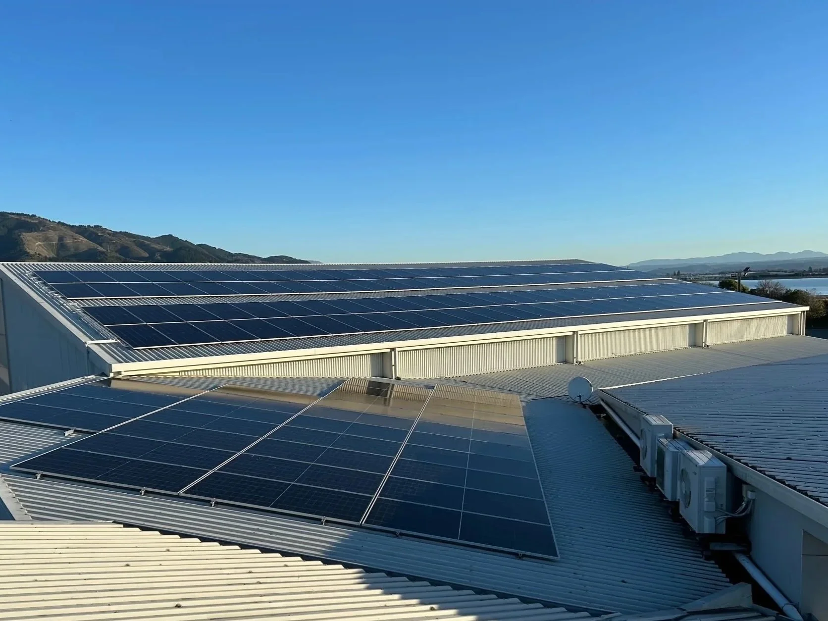 Rooftop solar panels installed on a metal building under a clear blue sky with mountains in the background.