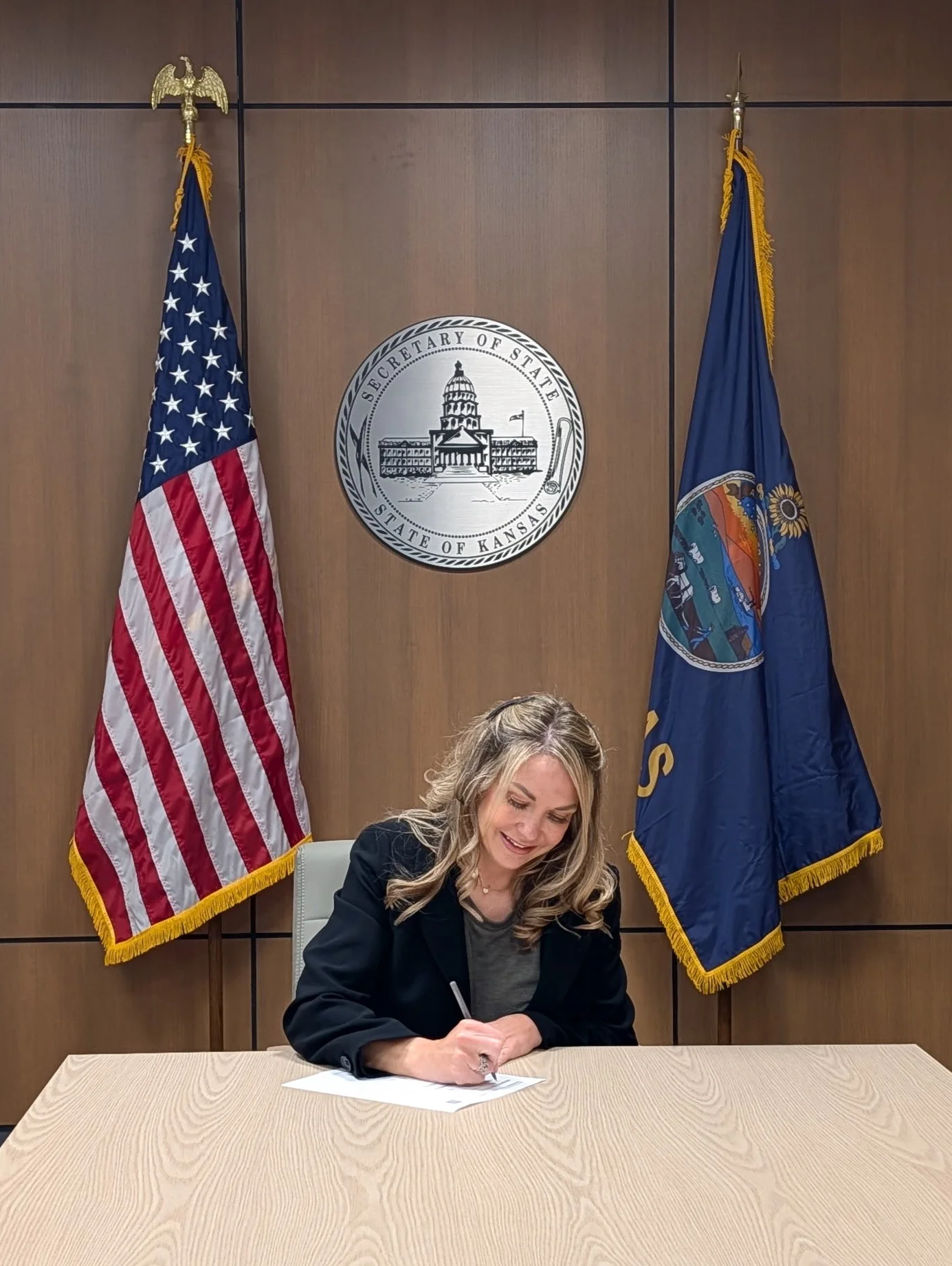 A woman with blonde hair, wearing a black blazer, sitting at a wooden table and signing a document. Behind her are two flags (the United States flag and the Kansas state flag) and a seal that reads 'Secretary of State Kansas'.