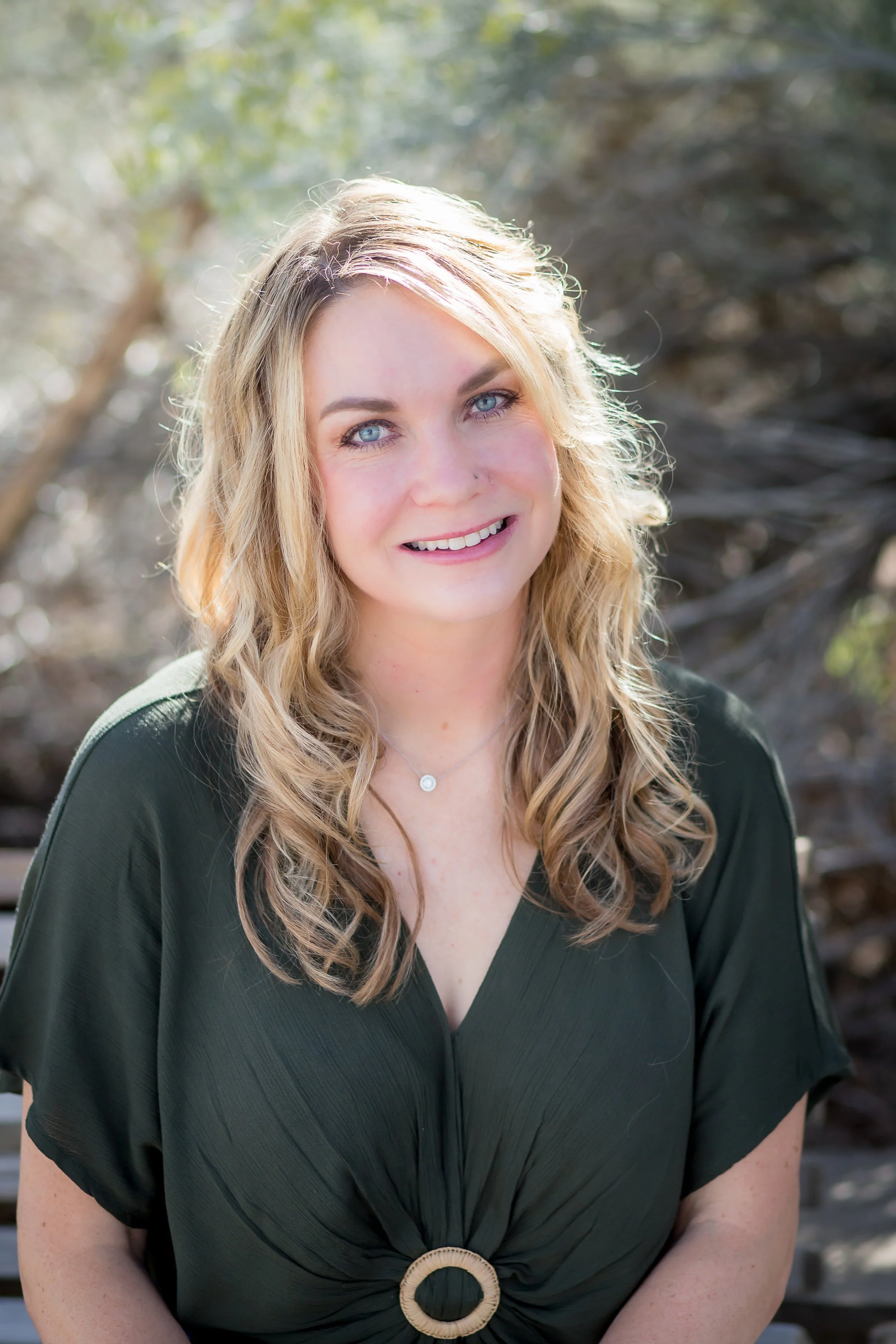 A young woman with blond wavy hair and blue eyes smiling outdoors with blurred trees in the background.