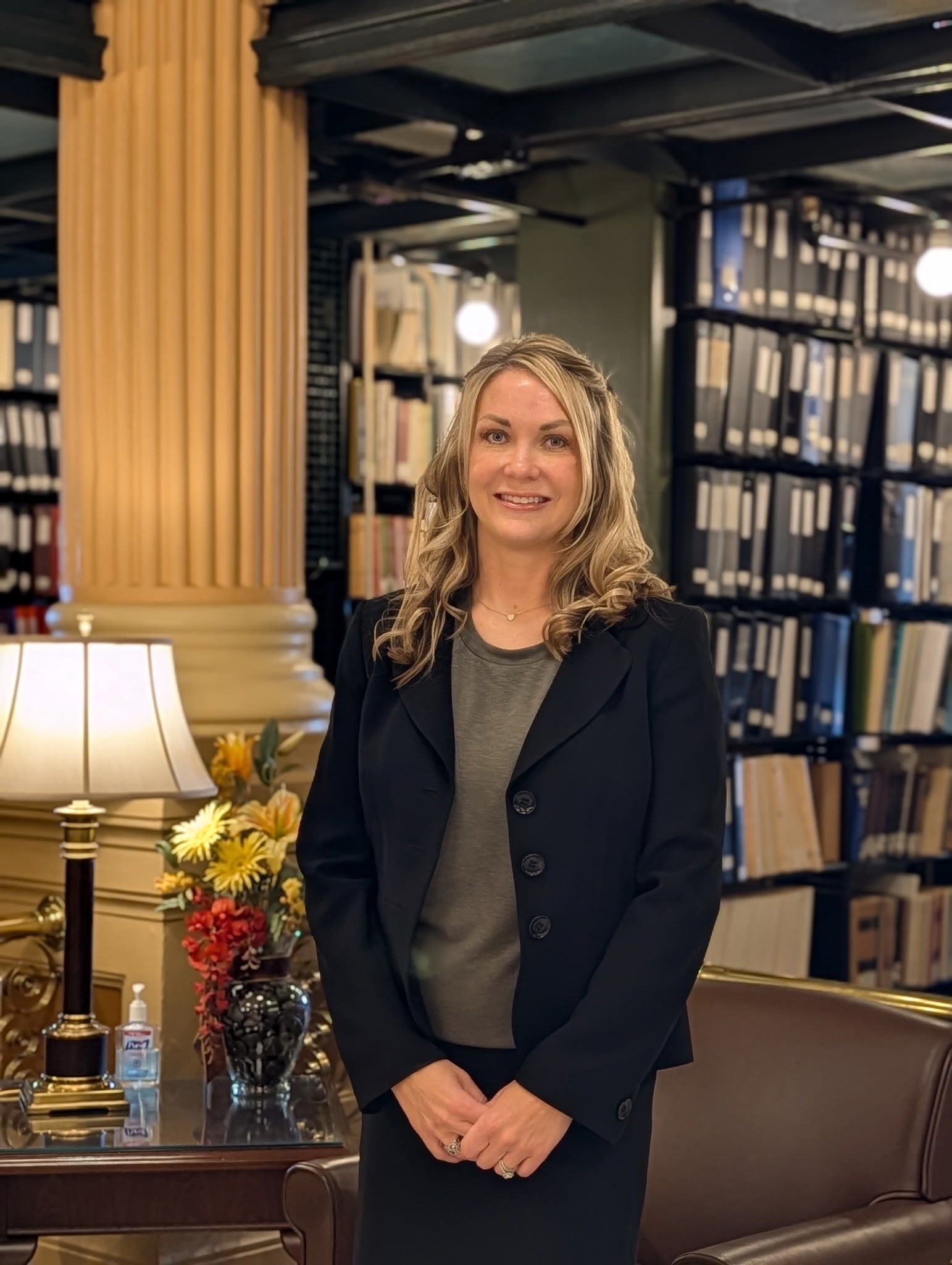 A woman in a black blazer standing in a room with bookshelves, a table with a floral arrangement, and a lamp.