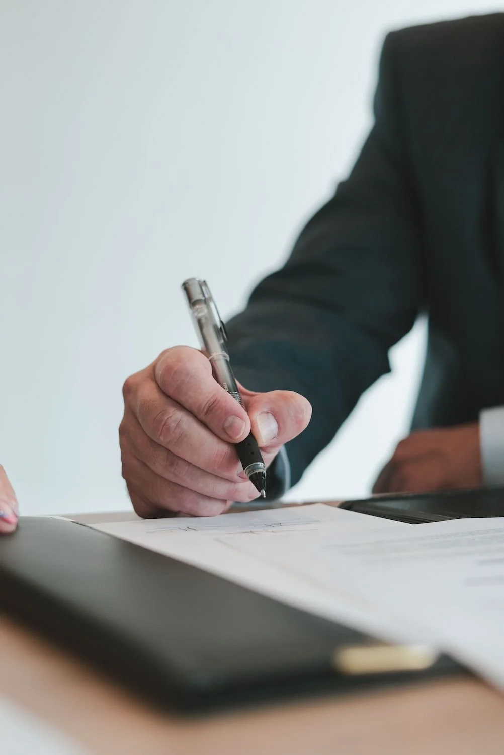 A person in a suit is signing a document with a black pen on a desk.
