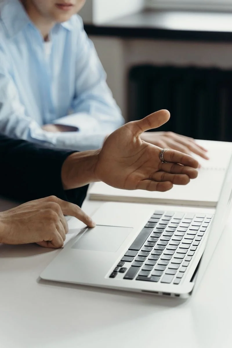 Two people discussing in front of a laptop, one gesturing towards the screen.