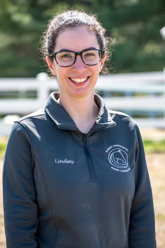 A young woman smiling outdoors, wearing glasses and a navy blue zip-up jacket with a logo that reads "Leddy Therapeutic Riding Center" and the name "Lindsey" embroidered on it.