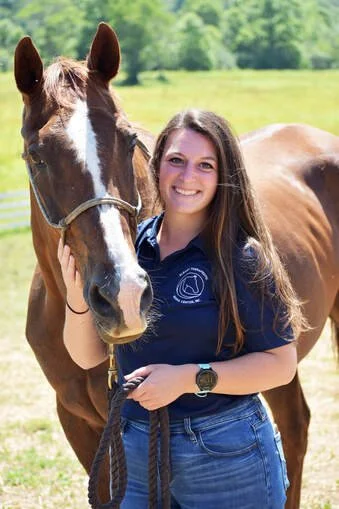A young woman with long hair smiling and holding a horse's bridle outdoors in a green field.