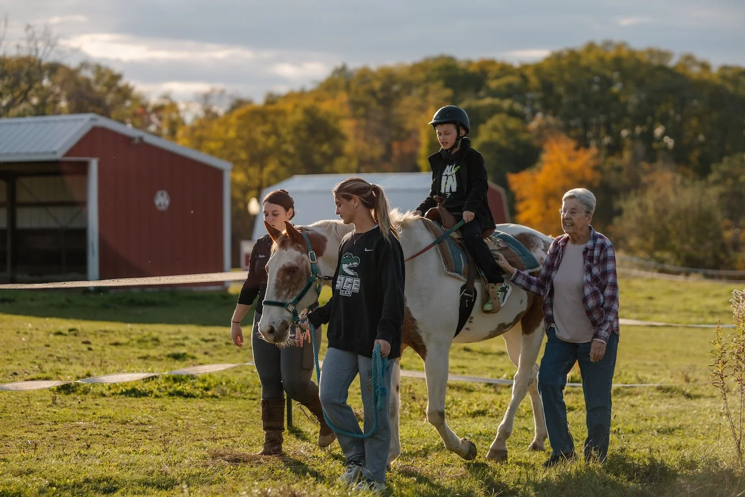 People helping a young girl ride a white horse with brown spots in a fenced field during daytime with trees and barn buildings in the background.