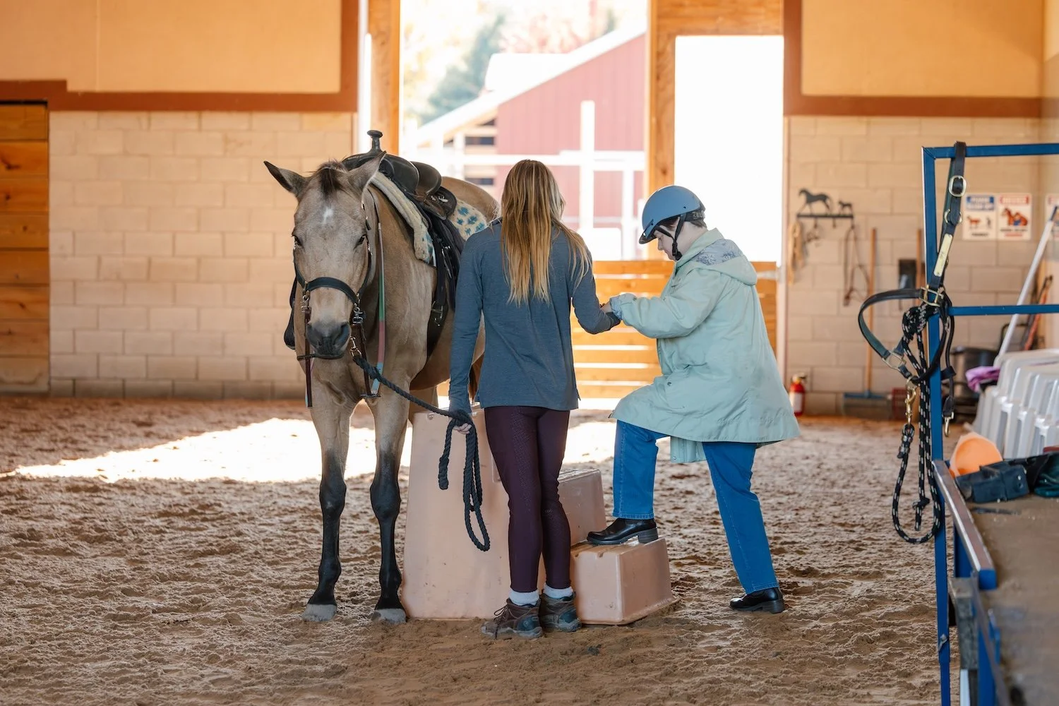 A woman in riding attire and a person in a helmet and coat are inside a horse stable, standing on step stools, and shaking hands over a saddle on a horse.