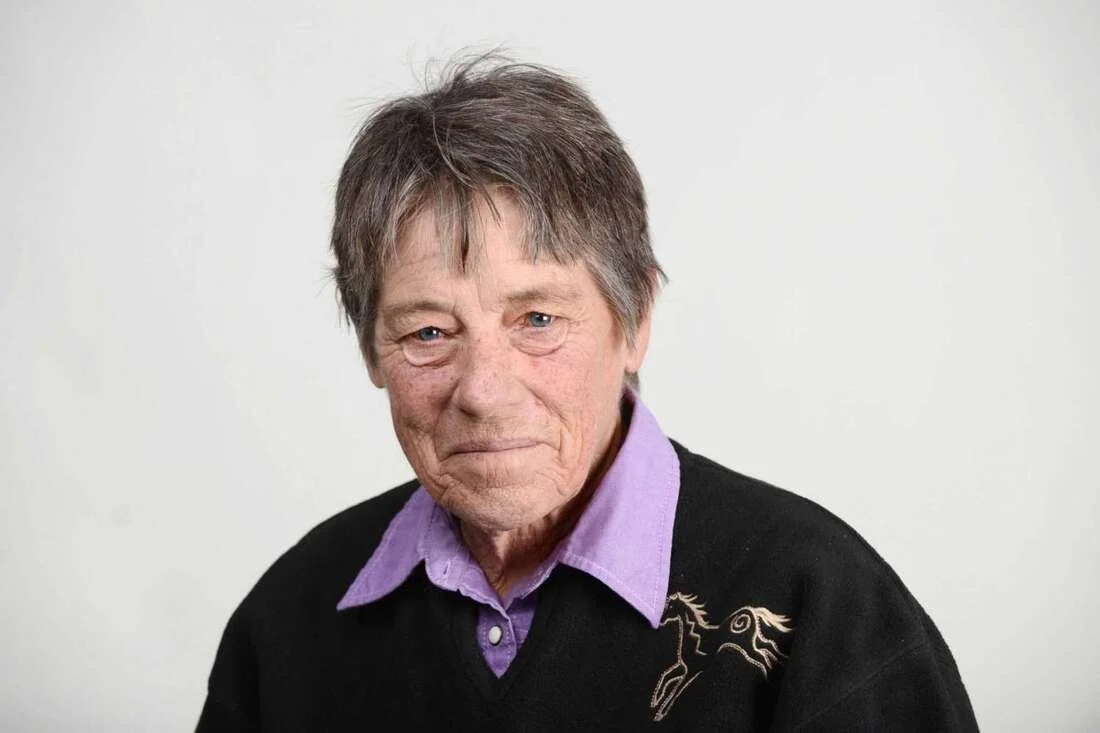 Portrait of an elderly woman with short gray hair, wearing a purple collared shirt and a black sweater with a horse embroidery, standing against a plain white background.