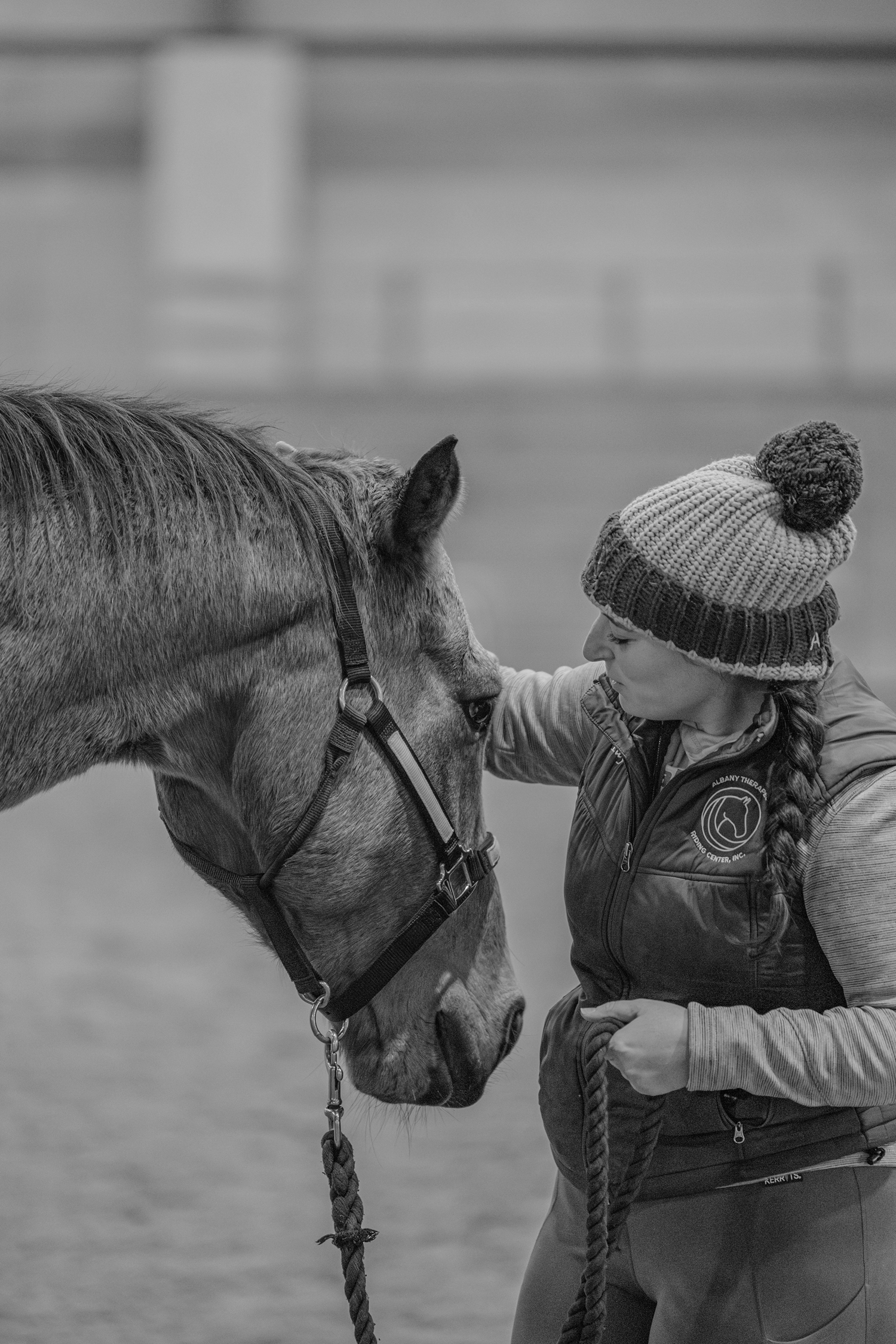 A young girl with braided hair, wearing a winter hat and jacket, gently touching the face of a horse indoors.