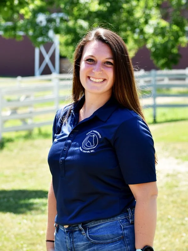 Smiling woman with long brown hair wearing a navy blue polo shirt with a logo on the chest, standing outdoors with green trees, a white fence, and a barn in the background.