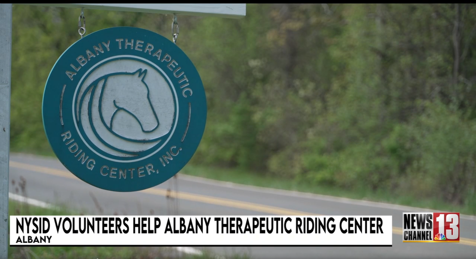 Blue circular sign with white outline of a horse's head, reading 'Albany Therapeutic Riding Center, Inc.' hanging on a chain outside.