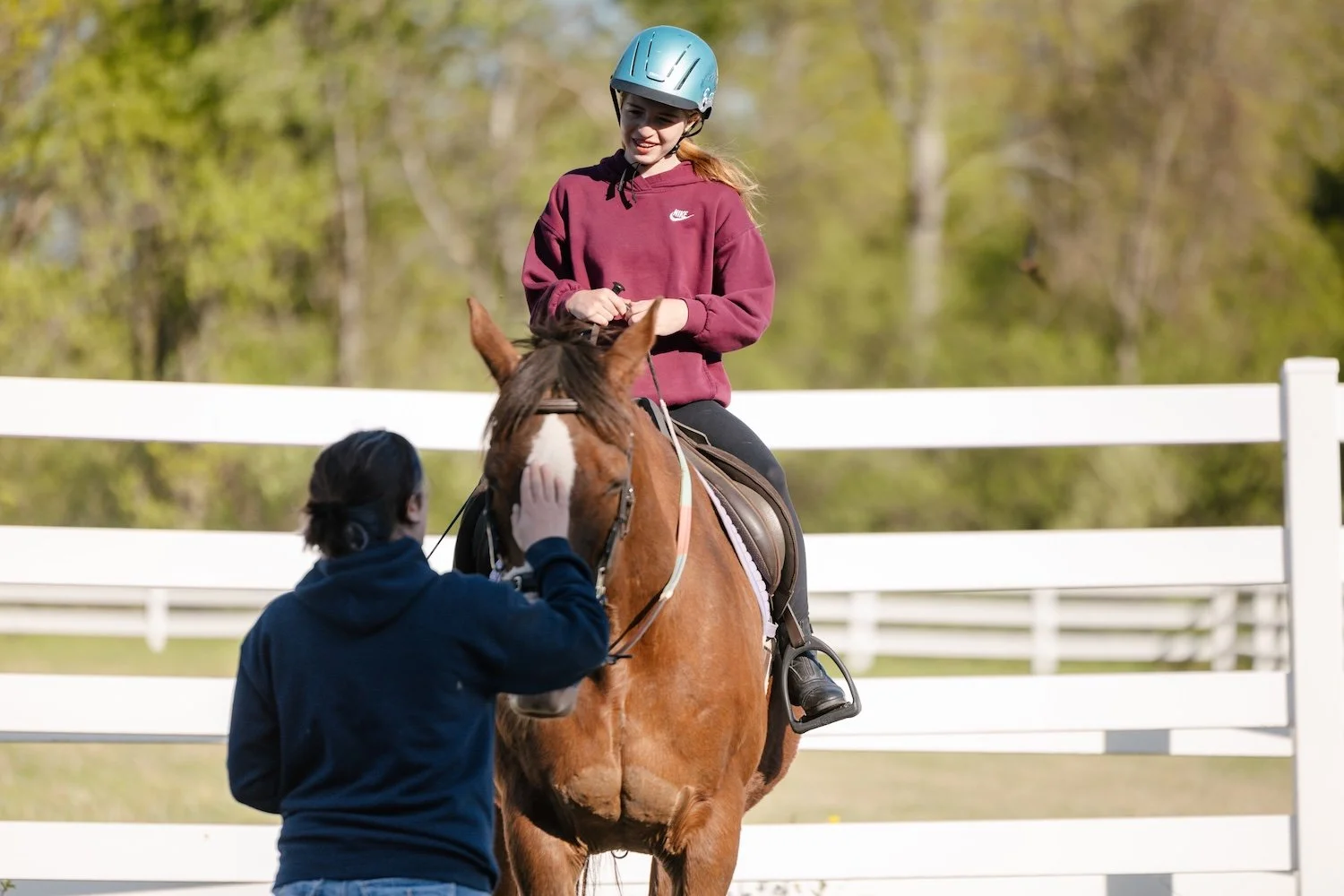 A young girl with a blue helmet riding a brown horse in an outdoor riding arena, while an instructor or assistant in a dark hoodie adjusts the horse's bridle.