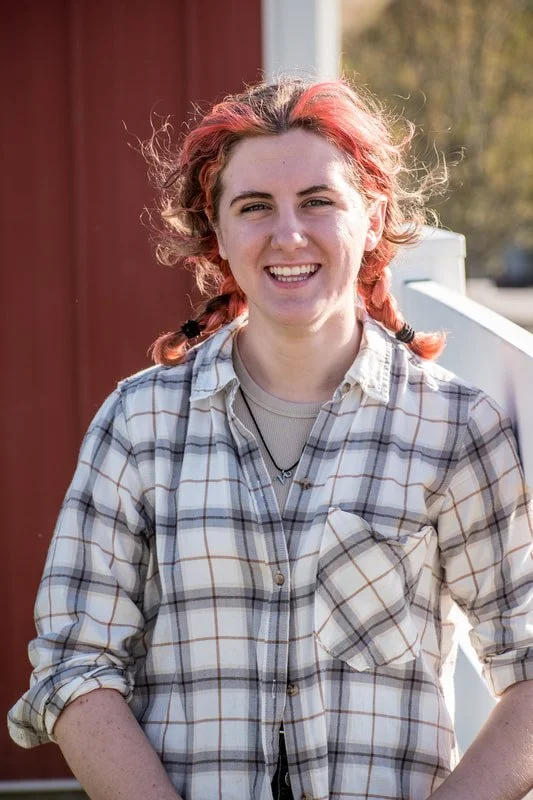 A young woman with red hair in braids smiling outdoors, wearing a plaid shirt over a light-colored t-shirt.