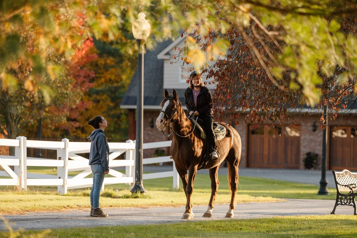 A woman riding a horse on a path in a park, with another woman standing nearby. The scene is framed by colorful autumn leaves and trees, with a white fence, a house, and a park bench in the background.