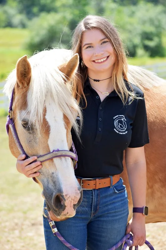 A young woman with long light brown hair smiling and holding a palomino horse by the bridle outdoors with green trees and grass in the background.