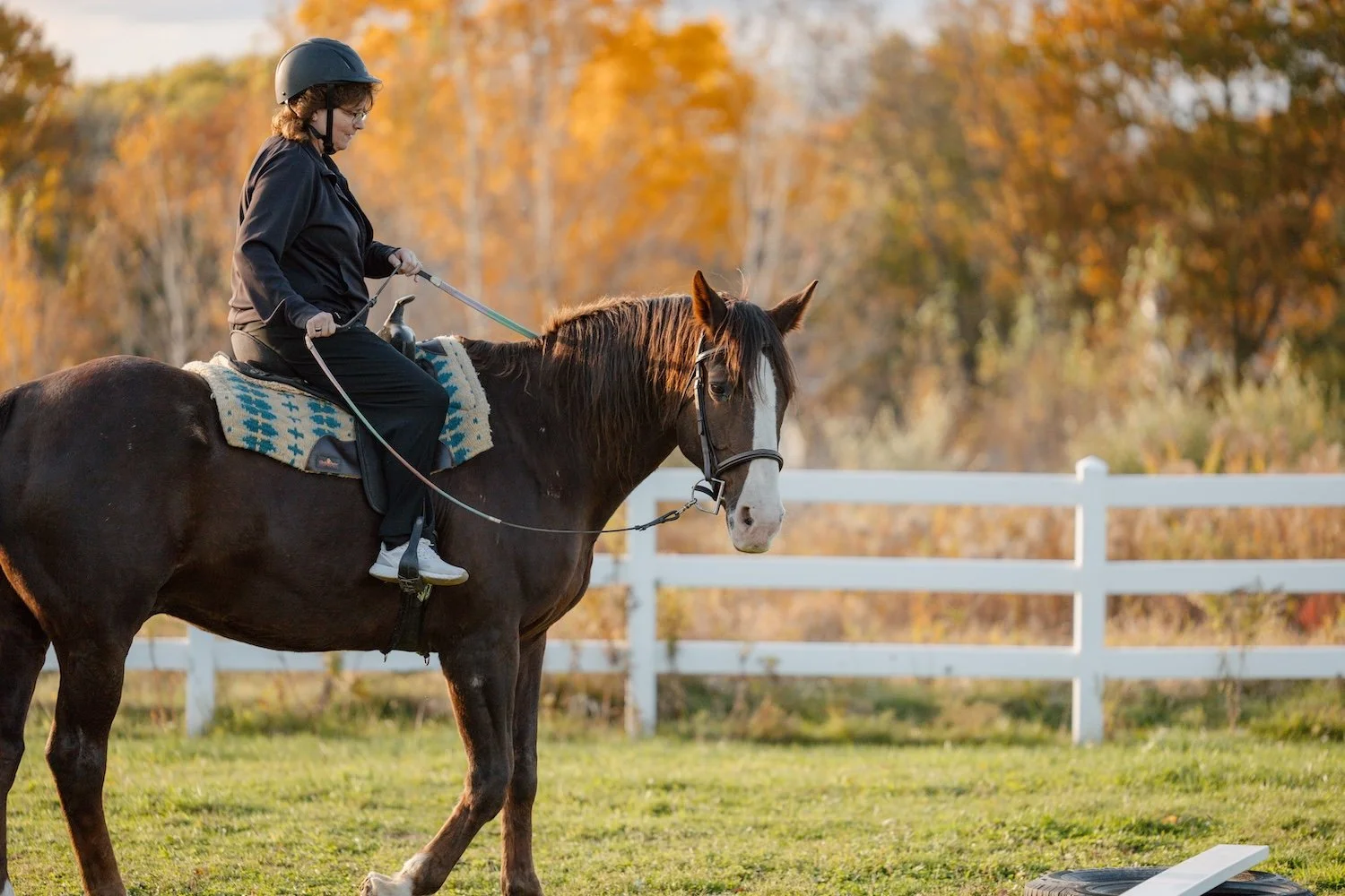 A person riding a brown horse with a white face mark in a fenced outdoor area during autumn, with trees in orange and yellow foliage in the background.