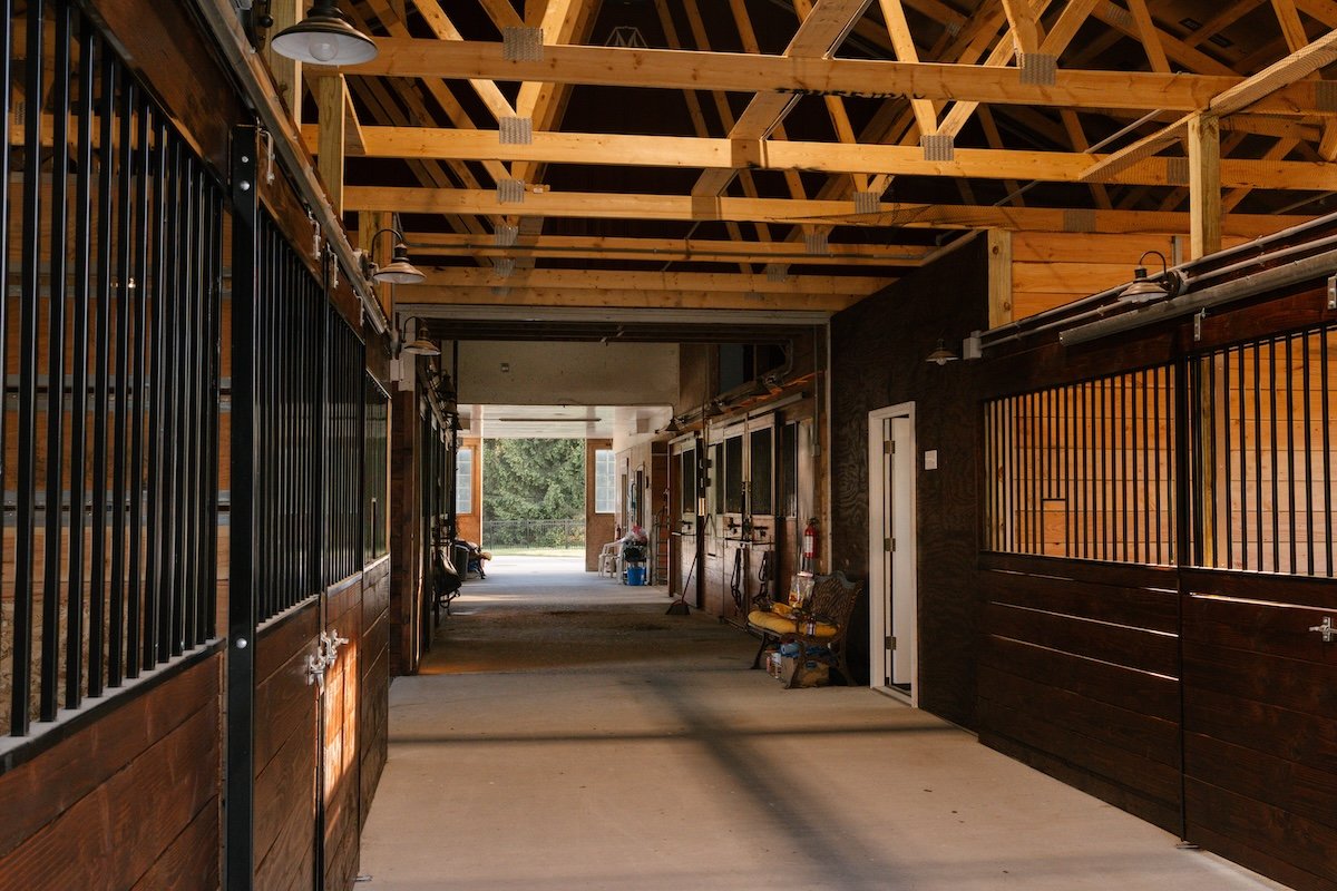 The inside of a horse stable with wooden walls and stalls on both sides, open at the end to an outdoor area with trees.