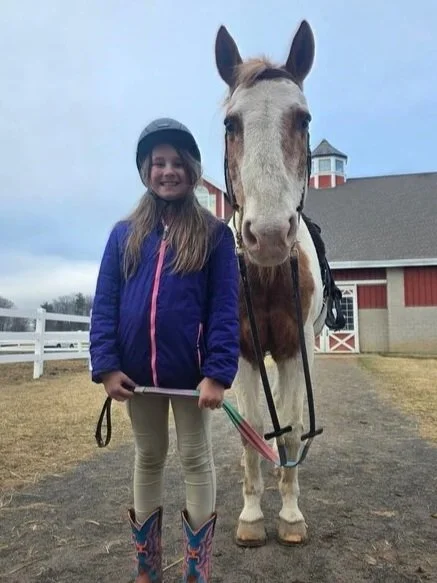 A girl wearing a helmet and purple jacket standing beside a white and brown horse with a barn in the background.