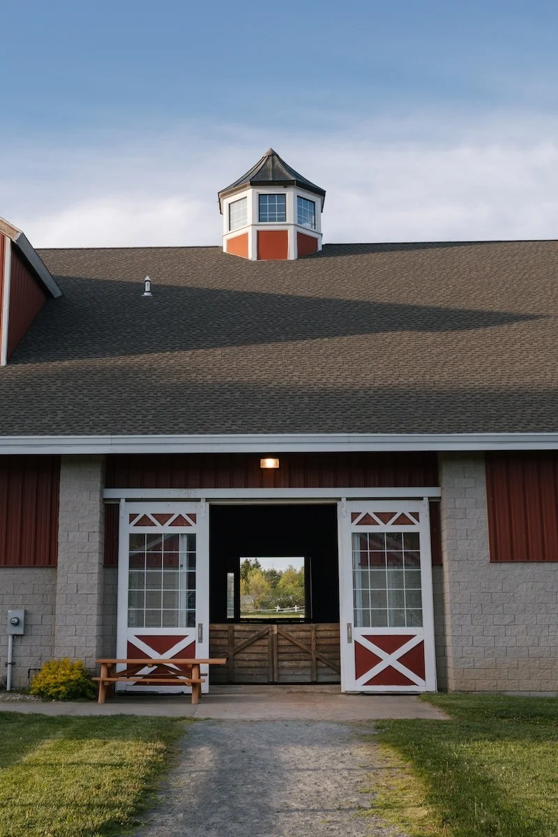 A red barn with white trim and a large open entrance, a gravel driveway, and a small wooden bench outside. The barn has a cupola on the roof and the sky is partly cloudy.