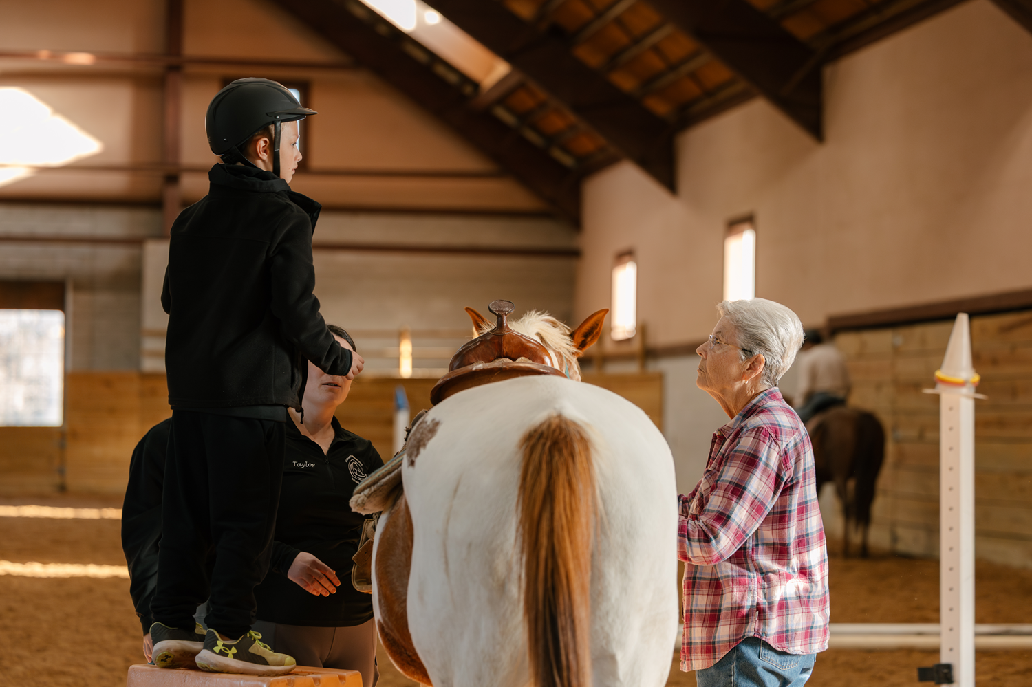 A young girl in a black helmet and jacket standing on a platform, preparing to ride a horse with assistance from two women in a stable, with sunlight streaming through the windows.