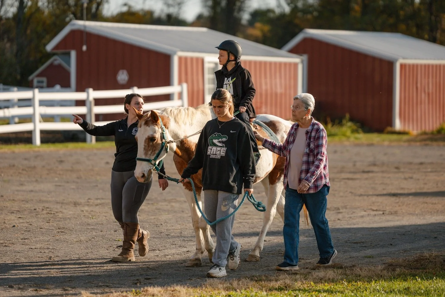 A young boy in a helmet riding a white and brown horse on a farm, accompanied by three women guiding and supporting him.