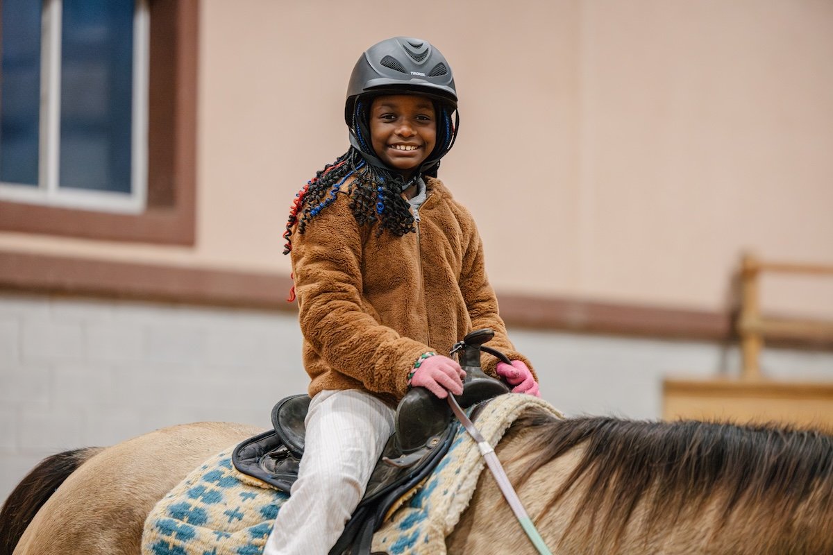 Smiling young girl with braids wearing a brown jacket and pink gloves riding a light brown pony inside a stable or riding area.