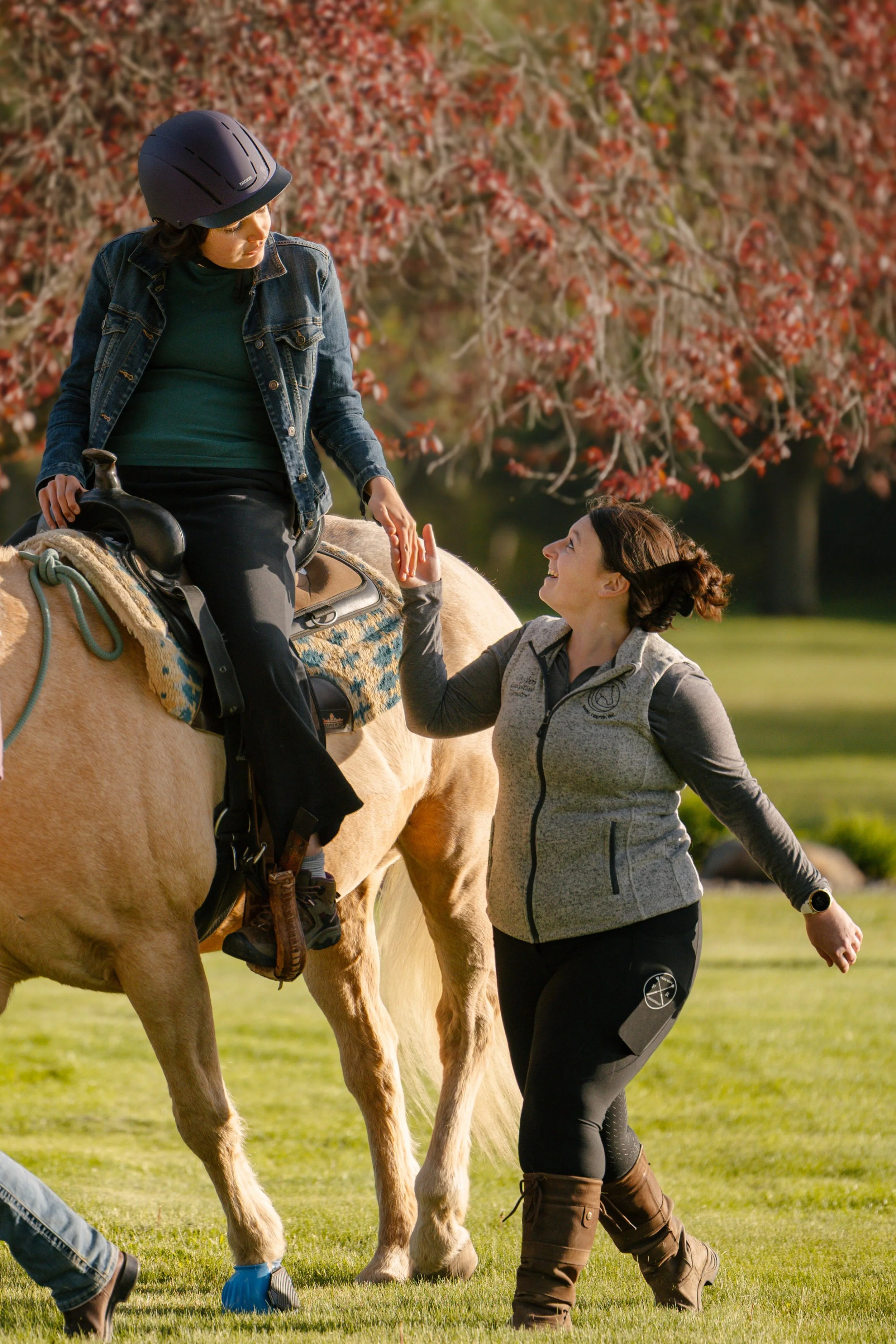 A woman dressed in casual riding gear is sitting on a light-colored horse, holding the reins. She is wearing a black helmet, denim jacket, and dark pants. Another woman, standing nearby in outdoor attire with a vest and black pants, is reaching out and smiling, touching the rider's hand. The background features trees with red leaves, suggesting fall, and the grass is green.