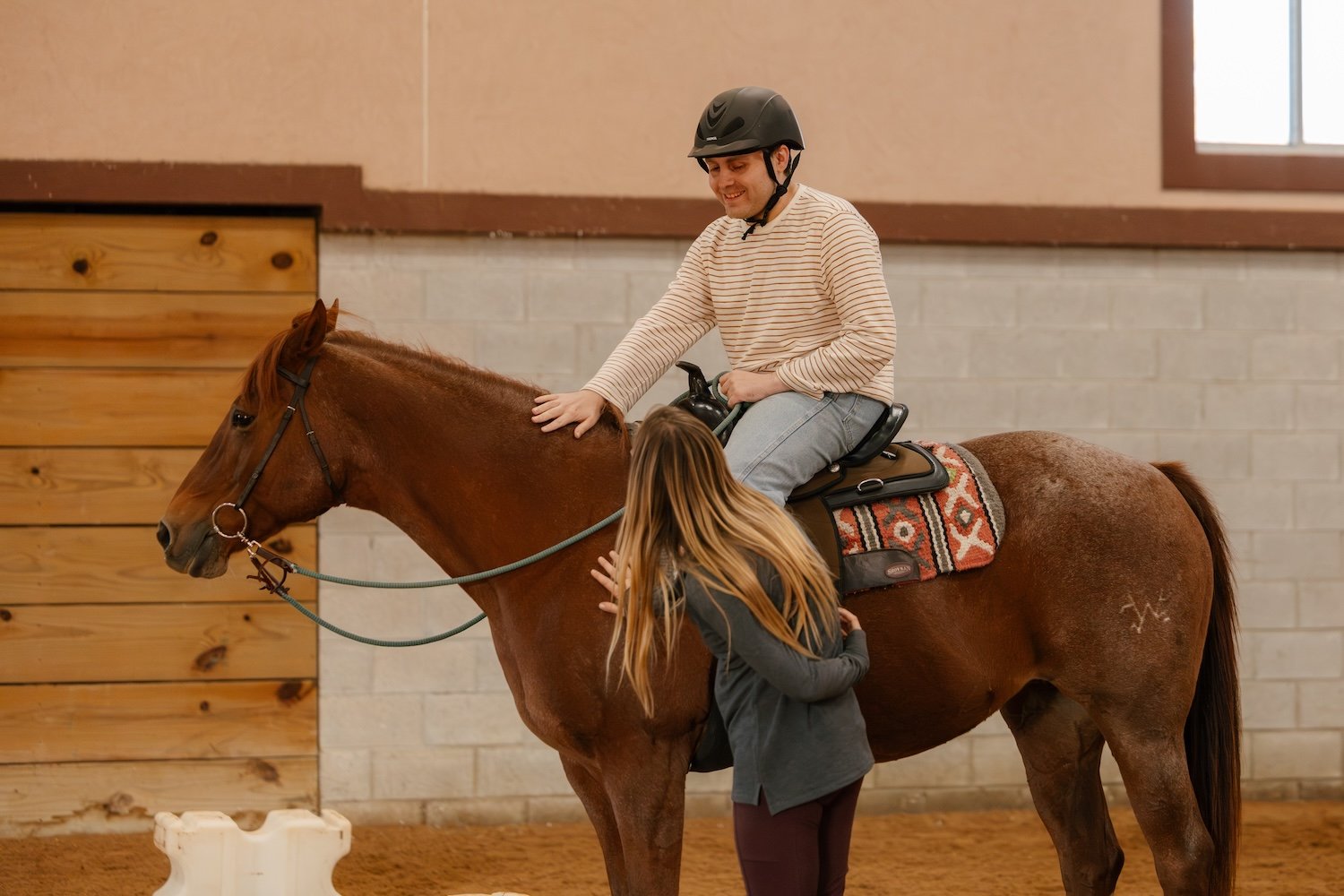 A man wearing a helmet and striped shirt riding a brown horse inside a riding arena, being assisted by a woman with long hair helping the horse.