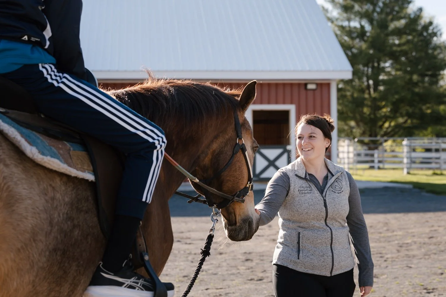 A woman smiling and holding a horse's halter while a person riding the horse is partially visible. They are in an outdoor riding area with a barn in the background.