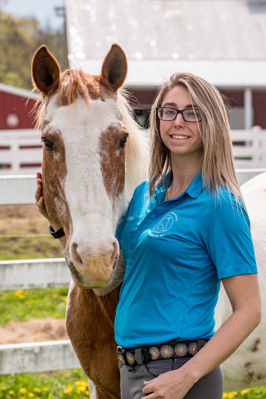 A woman in a blue polo shirt standing next to a white horse with brown patches, both looking at the camera, in a farm setting.