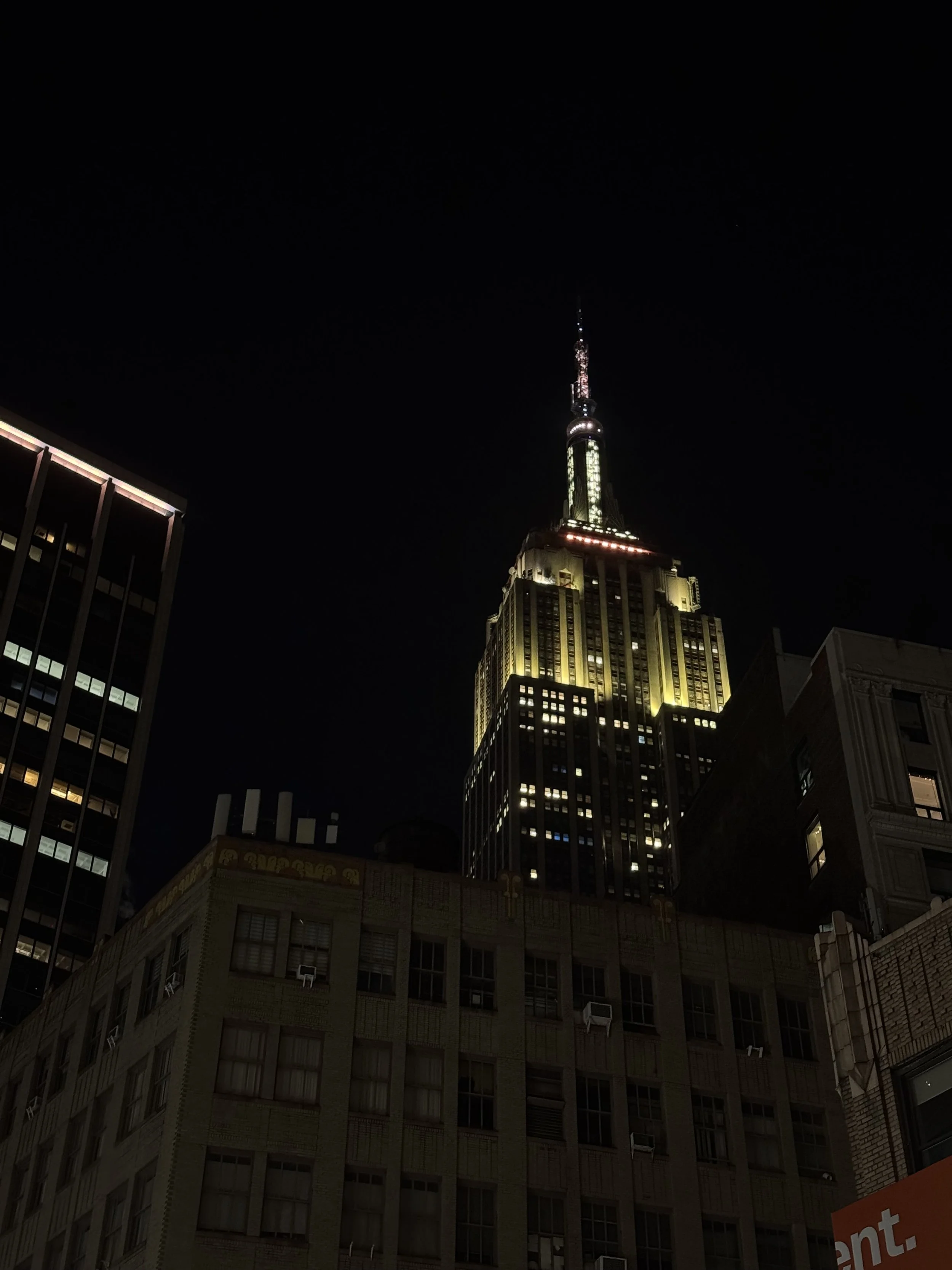 Nighttime view of the Empire State Building illuminated with lights, with surrounding buildings visible in the foreground.