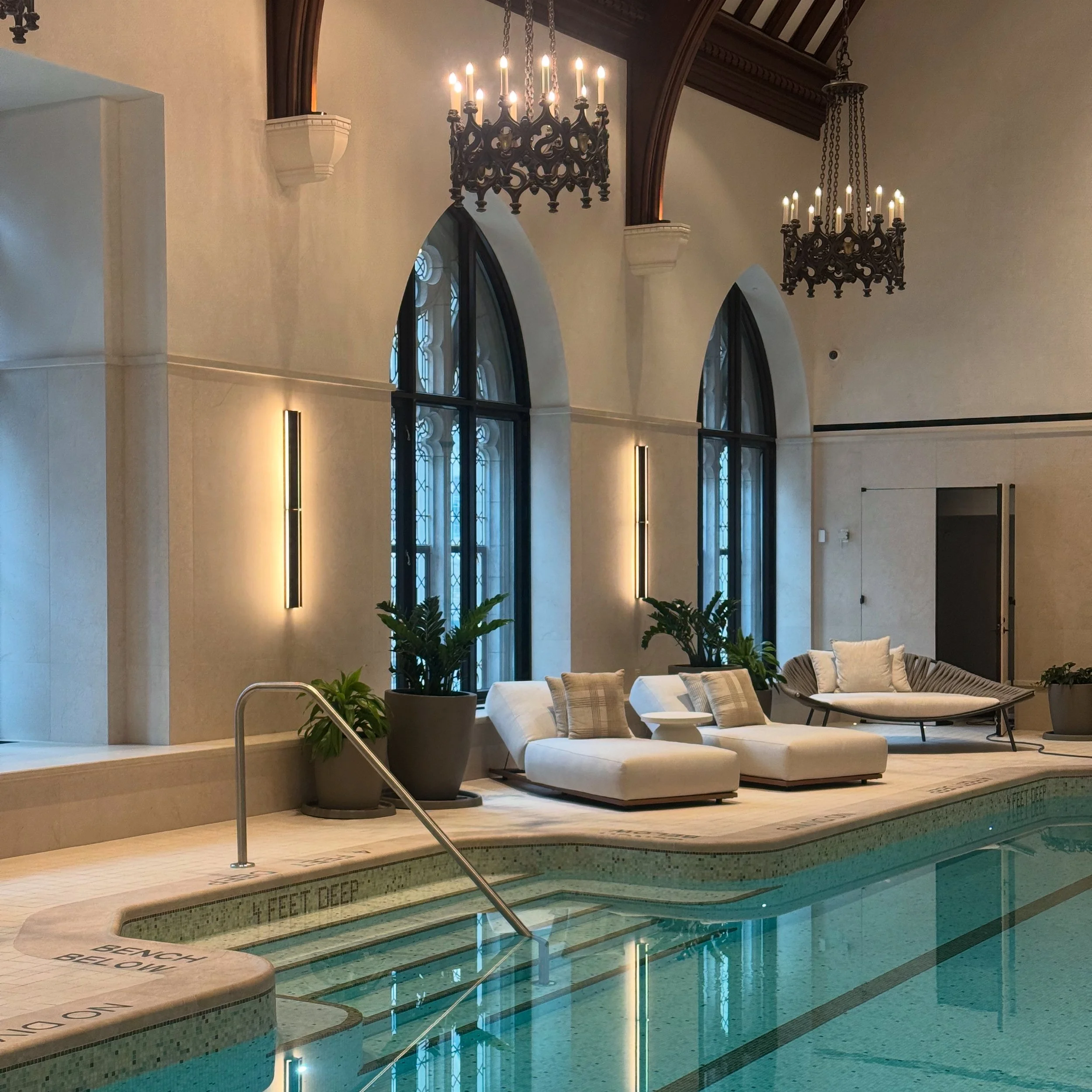 Indoor swimming pool area with a shallow end marked by a line on the floor, surrounded by lounge chairs and potted plants, with large arched windows and elegant chandeliers hanging from a high ceiling.