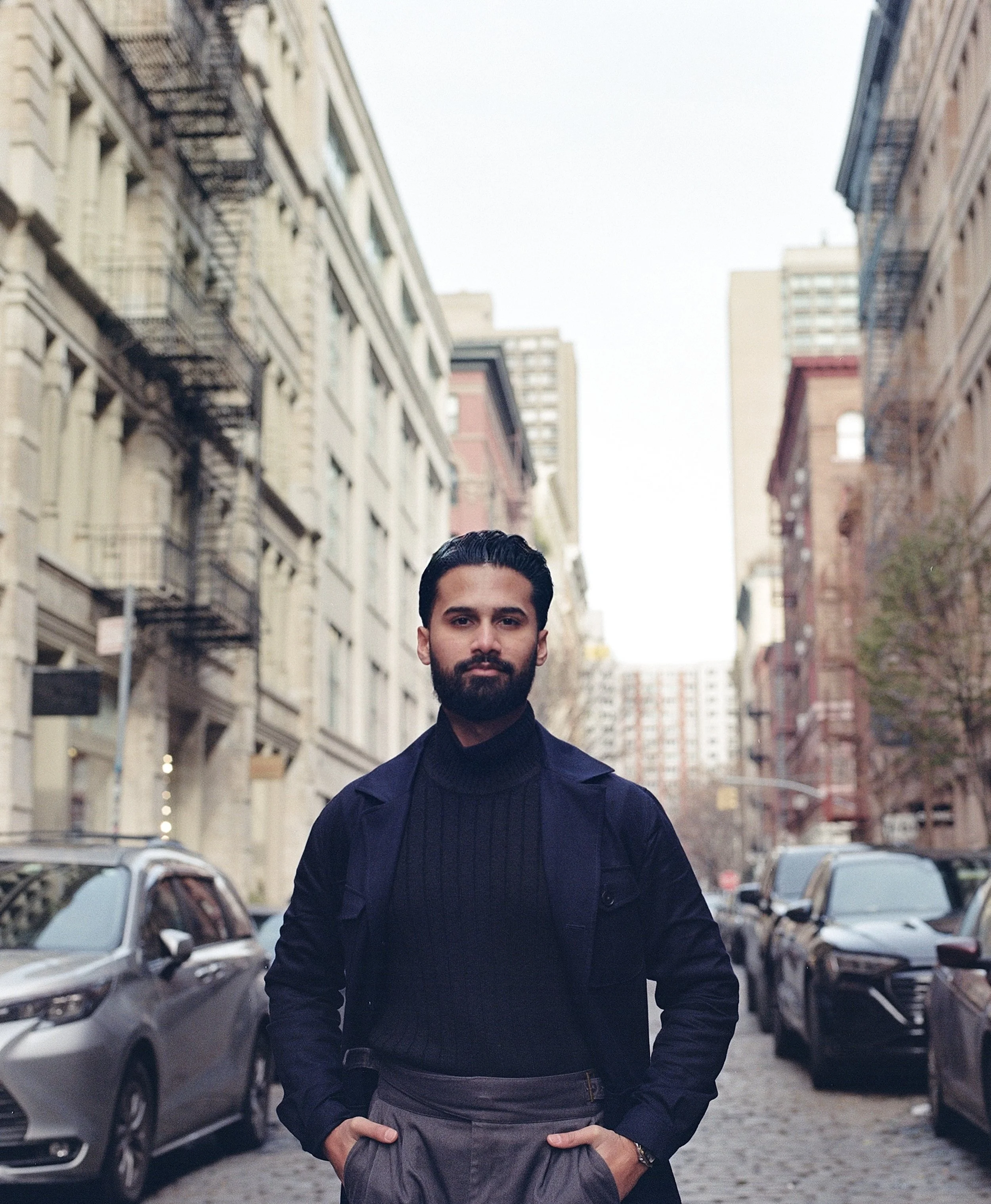 A man with dark hair and a beard standing on a city street with buildings and parked cars in the background. He is wearing a dark jacket and gray pants with his hands in his pockets.