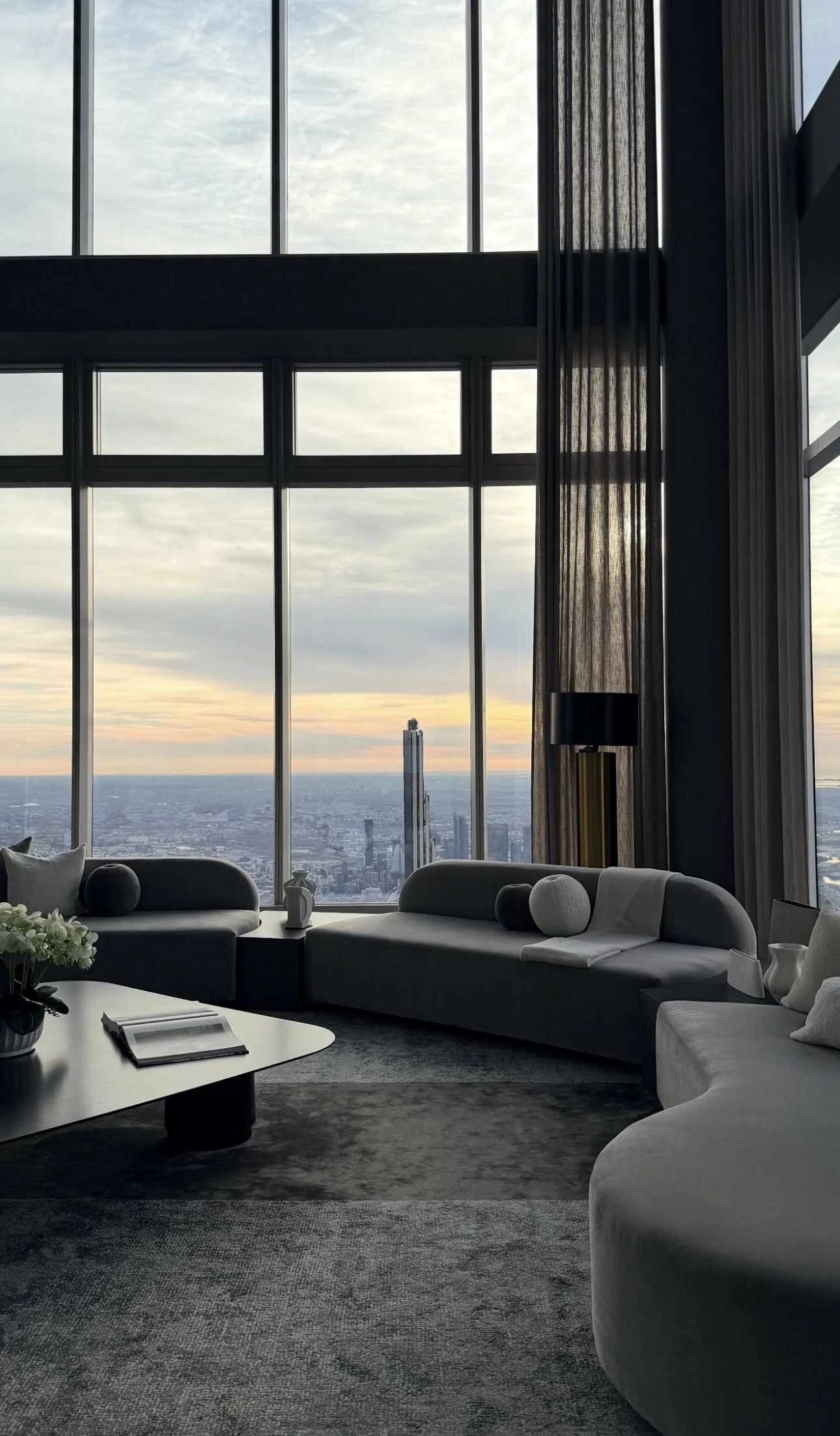 Living room in a high-rise apartment with large windows showing a cityscape sunset view, modern sofas, a coffee table, and decorative pillows.
