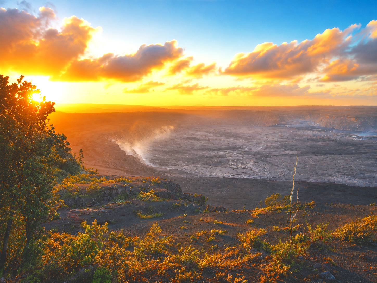 Volcanic crater at sunset with smoke rising from the lava, surrounded by green vegetation and a cloudy sky.
