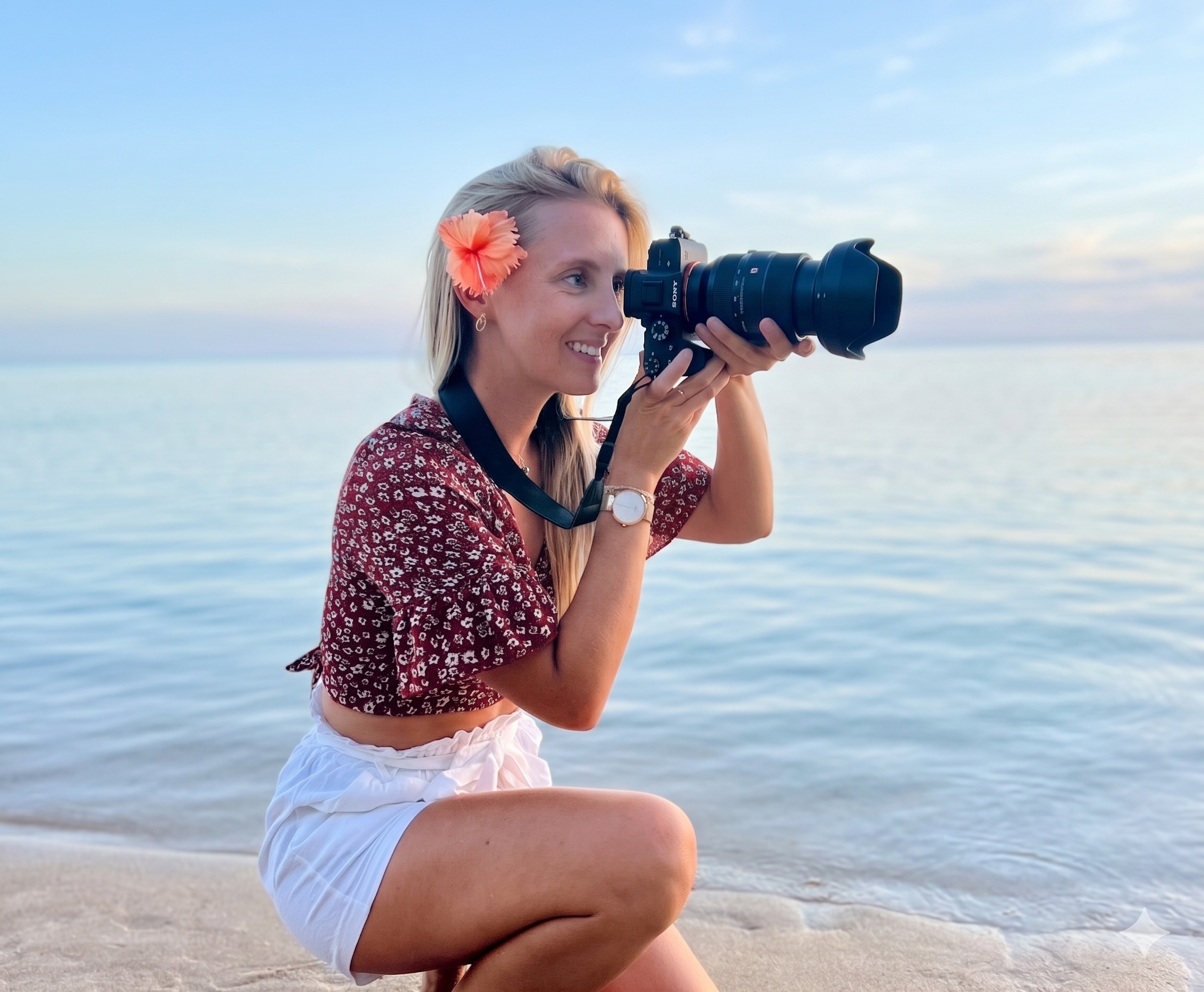 Une femme assise sur la plage, tenant un appareil photo grand angle en train de prendre une photo, avec un bandeau de fleurs dans ses cheveux, un ciel dégagé en arrière-plan. Megane S Photography à Tahiti