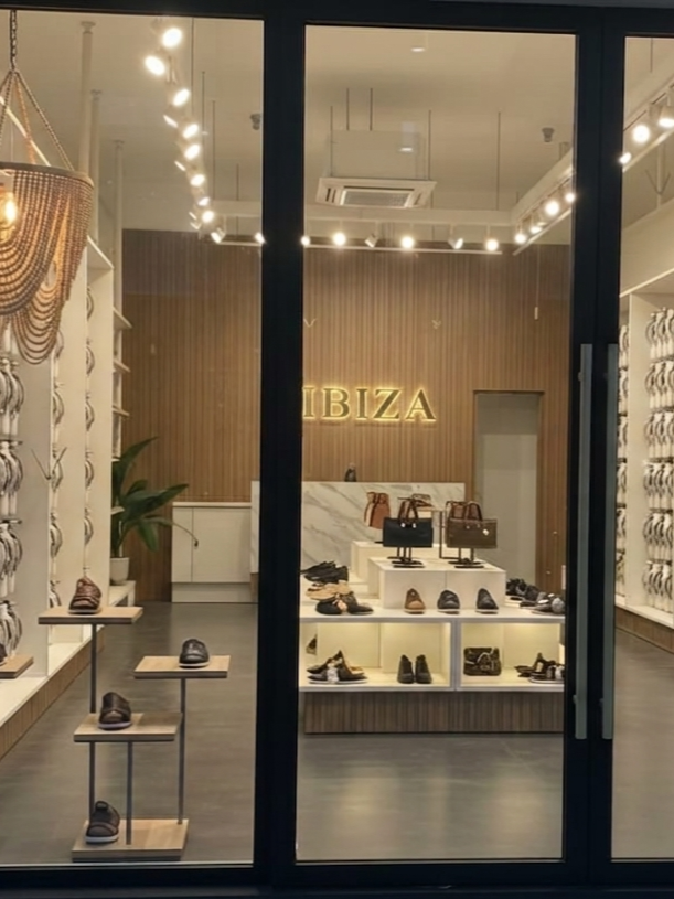 Vitrine d'une boutique de chaussures avec des chaussures et des sacs à main exposés, panneau lumineux indiquant 'IBIZA', intérieur moderne avec éclairage au plafond et murs en bois.